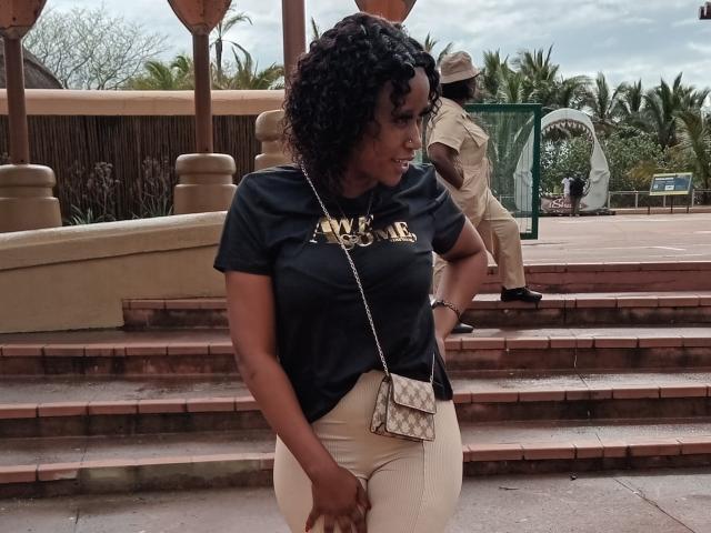 LisaLena with curly black hair, dark t-shirt, and white pants, holding a small purse, standing on brick steps, looking to the side, with a man in a tan uniform.