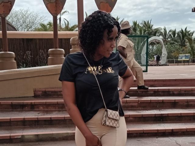 LisaLena with curly black hair, wearing a black "AMERICA" shirt, white pants, and a small quilted purse, stands on brick steps, looking down, with a security guard in the background.