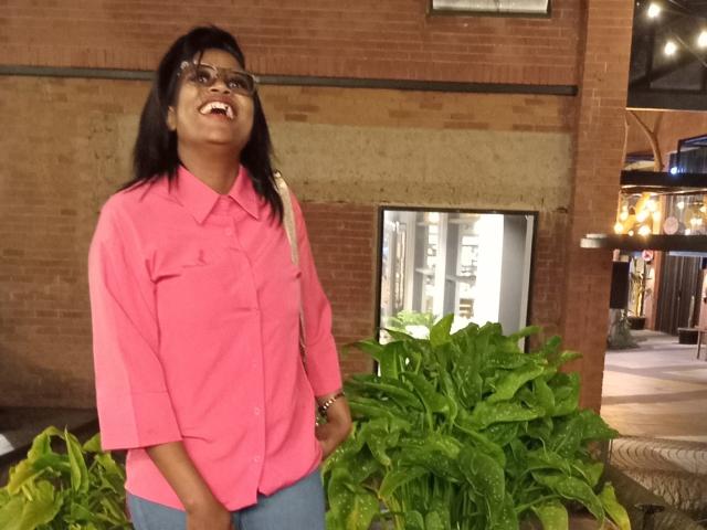 LisaLena with dark hair and glasses, wearing a pink shirt and blue jeans, smiling widely, standing in front of green leafy plants. She's got a cute, happy face.