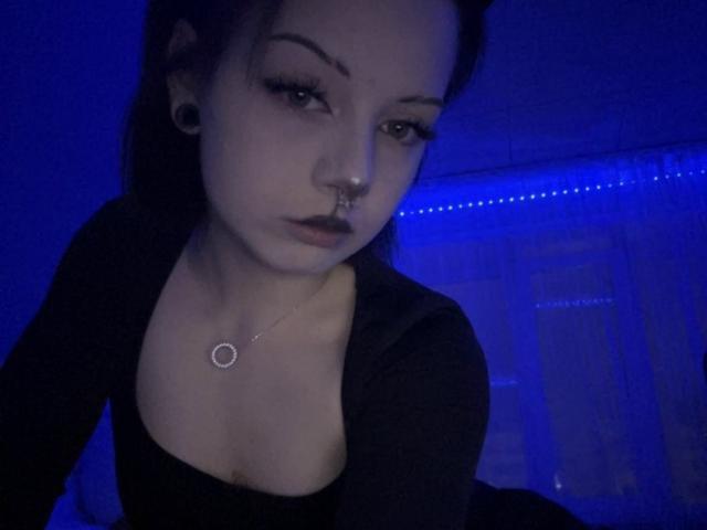 AnmiXHot with pale skin, dark lipstick, nose ring, black top, small tits, black hair, and earrings, looking at camera in blue-lit room.