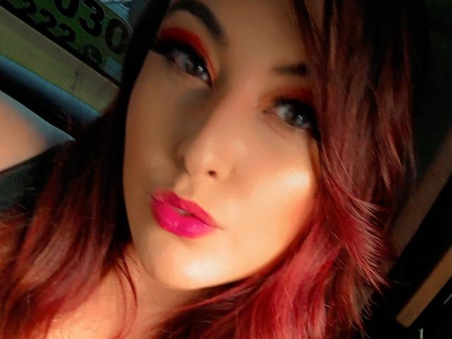 EmaRedFire ha l'eyeliner rosso, il rossetto rosa e i capelli con riflessi rossi in un selfie, con il sedere leggermente visibile sullo sfondo, e appare seducente con un leggero sorriso..