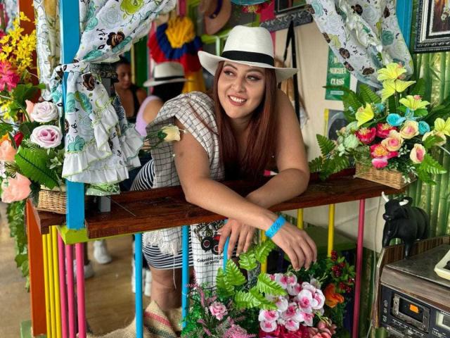 EmaRedFire with long brown hair, white hat, smiling, leaning on colorful wooden table, surrounded by flowers, wearing striped shirt, blue wristband, in vibrant, festive setting.