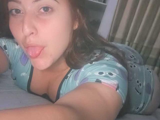 EmaRedFire, met haar tong uit, liggend op een bed, gekleed in blauwe pyjama's met smiley-gezichtjes, waarbij haar grote borsten zichtbaar zijn, en met bruin haar, op een licht onscherpe selfie..