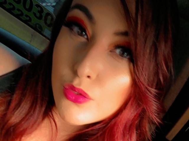 EmaRedFire a des lèvres rouges, des paupières roses et des cheveux rouge foncé, avec une légère moue et un regard doux, son visage illuminé par une lumière chaude..