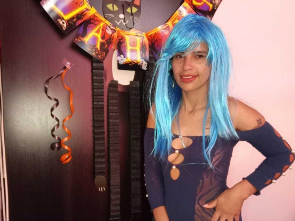 MileyHotty69 with blue wig, red lipstick, and dark off-shoulder top with cutouts, standing in front of a cat mask and garland, smiling confidently.