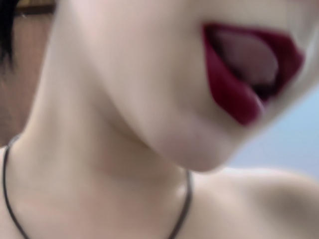 JanBetta's face, focused on her open mouth with red lipstick, showing her tongue. She has pale skin and wears a black necklace, with her neck and upper chest visible.