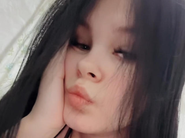 JanBetta with black hair, winking, pouting, and resting her hand on her cheek, showing soft pink lips and fair skin. Her eyes are half-closed in a cute, slightly shy look.