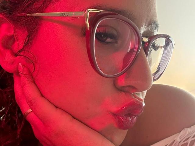 ValeryScot's face with red lighting, wearing big red-framed glasses, pouting lips, hand on cheek, curly brown hair, ring on finger, and a white off-shoulder top.