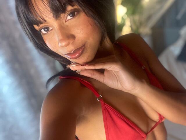 AylaCallowey with dark hair, wearing a red bikini top, showing her tits, hand near her face, smiling, brown eyes, tan skin, natural light, soft focus.