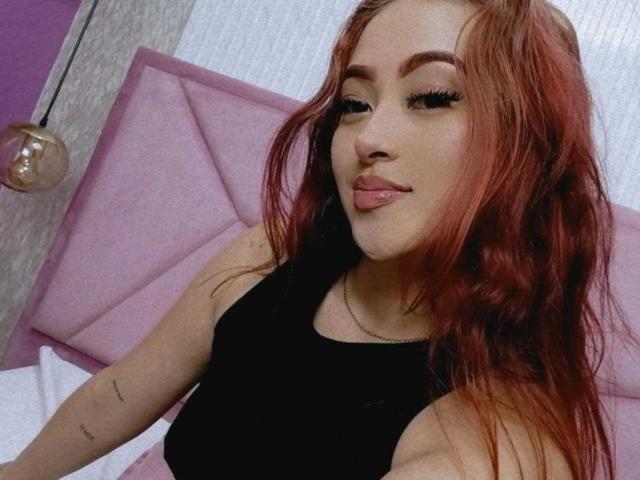 SamaraCuttie, com cabelo ruivo, camiseta preta e um sorriso discreto, exibindo seus seios com cílios longos, sentada em uma cama rosa, usando um colar de prata..