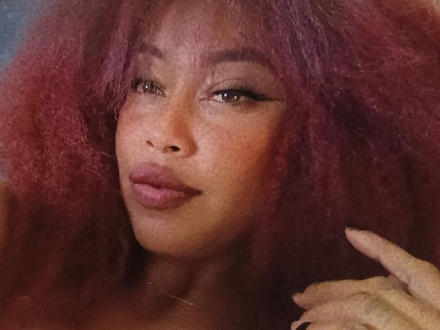 DalilaHeinz with dark brown skin, red curly hair, full pink lips, and subtle makeup, looking slightly to the side with a hand near her face, showing off her fingers.