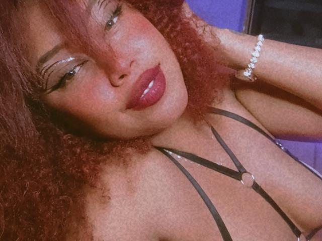 Dalila Heinz mit lockigem, rotem Haar, glänzenden, rosafarbenen Lippen und einem schwarzen, trägerlosen Top, das ihre Brüste zeigt und ein Armband an ihrem rechten Handgelenk trägt. Ihre Augen sind halb geschlossen..