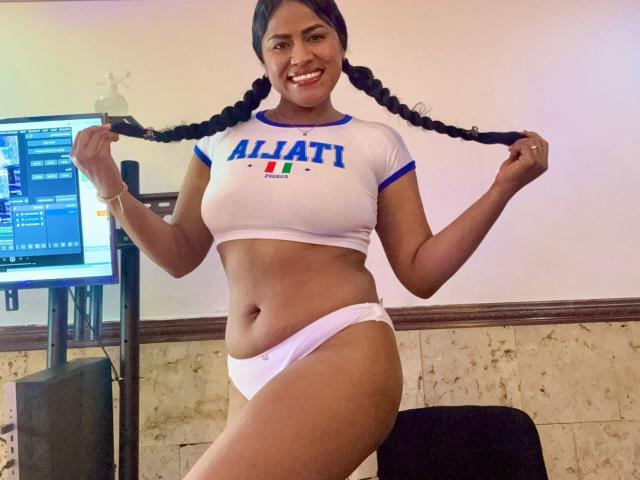 IvannaDuran with dark skin, smiling, holding braids, wearing white crop top and white panties, showing her tits and ass, standing in front of a computer.