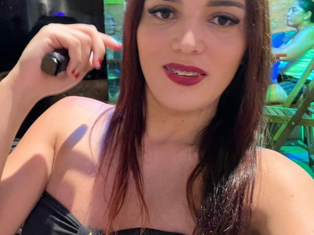 Close-up of Milenareistrans with long brown hair, fair skin, and dark red lipstick. She holds a black object in her right hand, index finger pointing at it. She has dark eyeliner and red-painted nails. She wears a strapless black top. The background is blurry, with green and blue hues. The image has a casual, slightly grainy texture. Her expression is neutral with a slight smile. The focus is on her face and upper body.