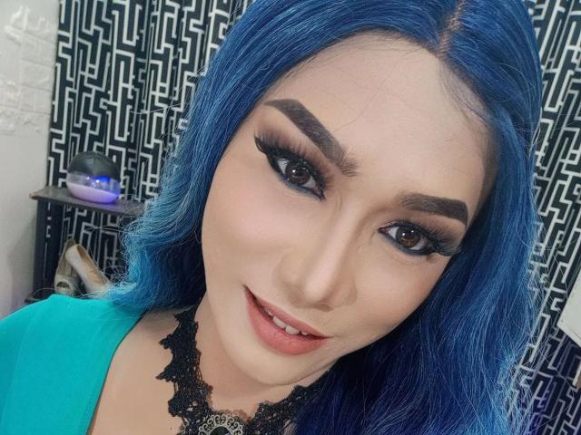 A close-up of HeavenlySelfsuck-hot with vibrant blue hair, thick black eyebrows, and long, dramatic black eyeliner. She has a light skin tone, full lips with a subtle pinkish-red lipstick, and a slight, confident smile. She's wearing a black lace choker and a turquoise top. The background has a black and white geometric pattern. Her makeup is bold and striking, contrasting with her bright hair. The image is sharp, emphasizing her facial features and makeup.