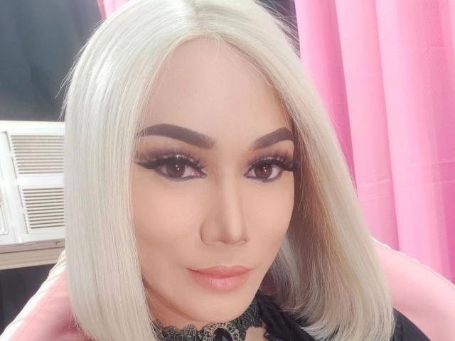 HeavenlySelfsuck - Uma mulher atraente com cabelo loiro platinado, sobrancelhas escuras, delineador marcante, cílios longos, batom rosa, colar de renda preta, sentada em uma cadeira rosa, com um aparelho de ar condicionado e uma cortina rosa ao fundo..