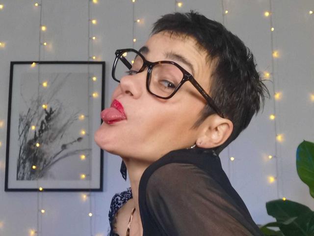 LaPhoenixElo with short black hair, glasses, red lipstick, tongue out, black sheer top, earrings, and a necklace, looking back with a playful expression.