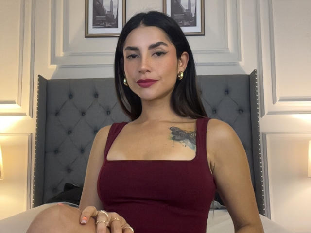 SofyaLane, avec ses cheveux foncés, son rouge à lèvres rouge et ses boucles d'oreilles dorées, portant un débardeur rouge, montrant ses seins, avec un tatouage sur la poitrine droite, assise sur un lit, regardant la caméra..