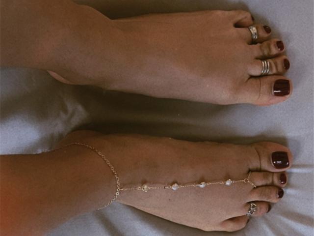 SofyaLane's feet, bare with dark red toenail polish, silver rings on toes, and a delicate ankle chain with small pearls on her right foot.
