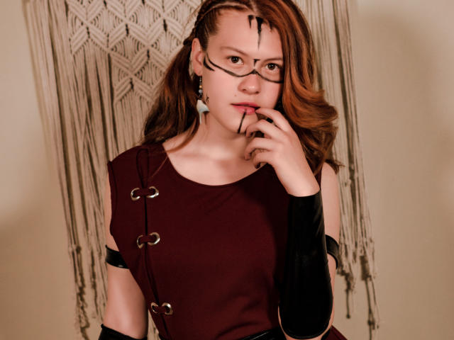 TaylorCarpert with glasses, brown wavy hair, red dress, black arm sleeve, finger on lip, earrings, looking at camera, beige background, soft lighting, sexy, thoughtful expression.