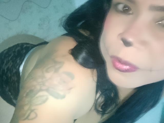 Chantali with shiny pink lips, black wavy hair, and a tattoo on her shoulder, wearing black lace panties, looking up at the camera. Her tits are slightly visible.