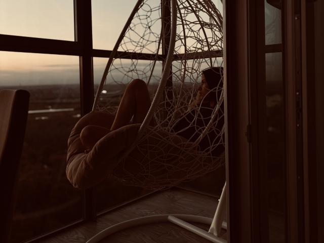 AngelaXSmile in a wicker hanging chair with her ass up, legs together, and arms around her head, silhouetted against a sunset through large windows. Dark, cozy, and sexy.