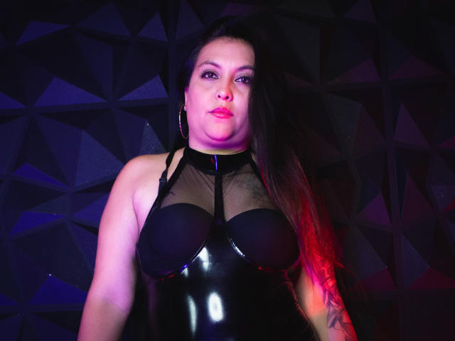 MissSusan with large boobs in a shiny black latex dress, sheer top, and long brown hair, wearing hoop earrings, in a purple and red lit photo.