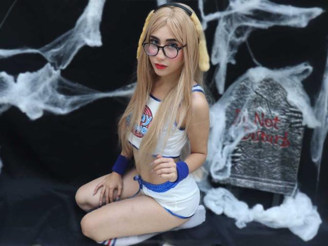 DinnyPink with long blonde hair, glasses, and yellow headphones, wearing a white and blue sports outfit, sitting with her legs crossed, showing her small ass and white socks.