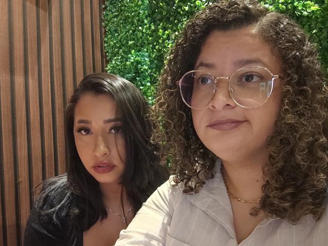 Marinomade with curly brown hair, glasses, and a white shirt, and Marinomade with black hair, red lipstick, and a black top, sitting together. Boobs, ass, tits not visible.