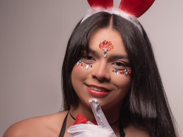 BabeCaty with dark hair, red bunny ears, white face paint, glitter, and a red gem on the forehead, smiling with a finger on her lips, wearing a black halter.