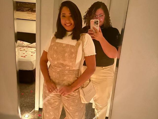 Marinomade with medium tits, wearing a pink floral dress over a white top, smiling in mirror selfie. Second Marinomade, curly-haired, in black shirt and white pants, takes photo.