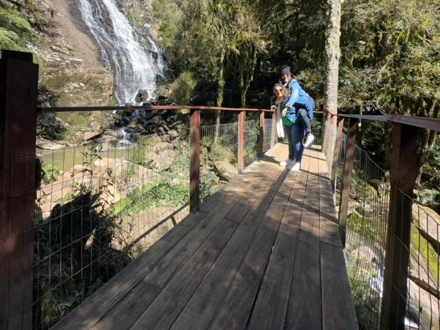 Marinomade is running on a wooden bridge with a red railing, wearing a blue jacket and green pants, waterfall in the background, sunlight filtering through trees.