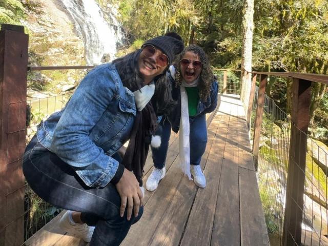 Marinomade with curly hair, wearing sunglasses, green shirt, and white scarf, stands crouched on wooden bridge. Marinomade with black hair, denim jacket, and grey scarf, crouches beside.