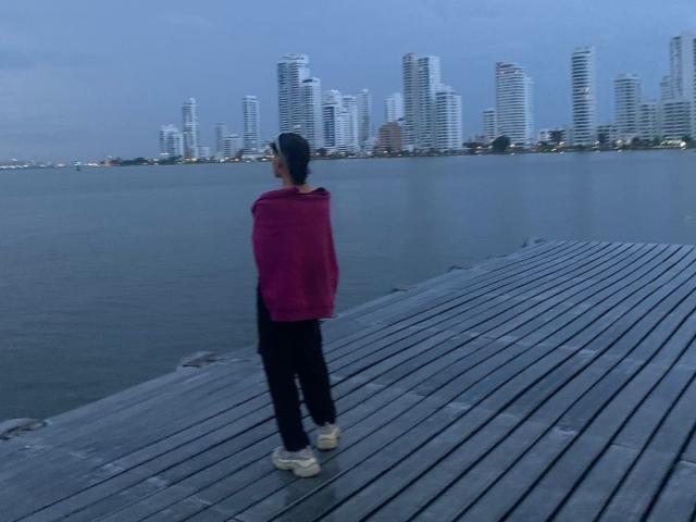 Angelitocute with a pink shawl, black pants, and white shoes, standing on a wooden dock, facing a city skyline of tall, white skyscrapers at dusk.