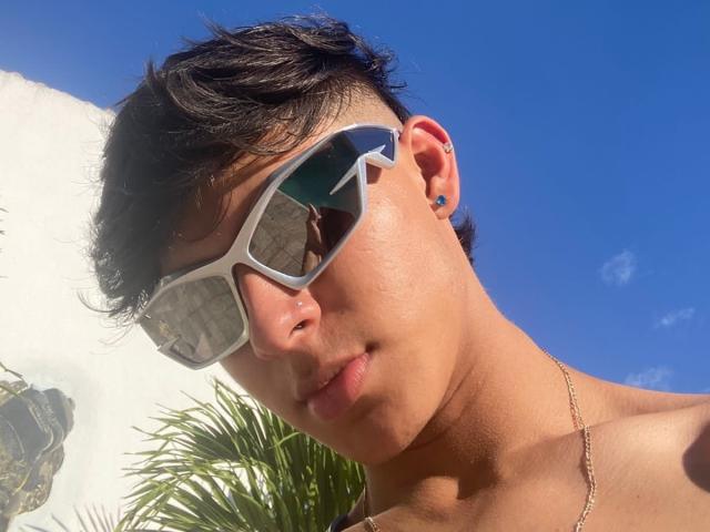 Angelitocute with shiny skin, short dark hair, and white sunglasses reflecting a blue sky, has a gold necklace and small ear piercings, looking slightly down.