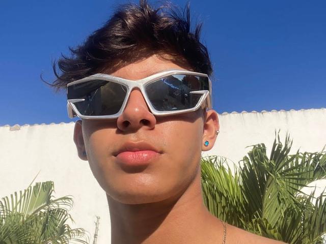 Angelitocute with short, dark, wavy hair, wearing white, geometric sunglasses, and turquoise earrings, has a cute, tan face and full pink lips, against a bright blue sky.