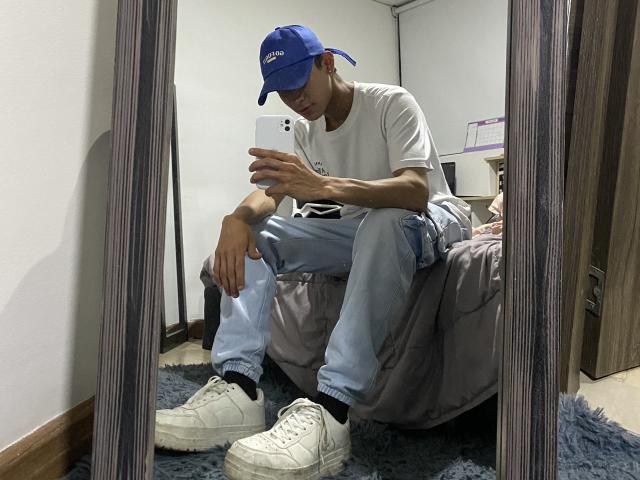 Mirror selfie of Angelitocute sitting on bed, wearing blue cap, white tee, light jeans, white socks, and white sneakers, holding phone, looking down, in dimly lit room.