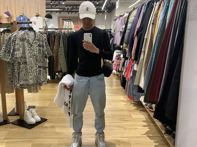 Angelitocute in a store, wearing a white cap, black long-sleeve, light blue jeans, and silver sneakers, holding a white shirt, standing in front of a clothing rack.