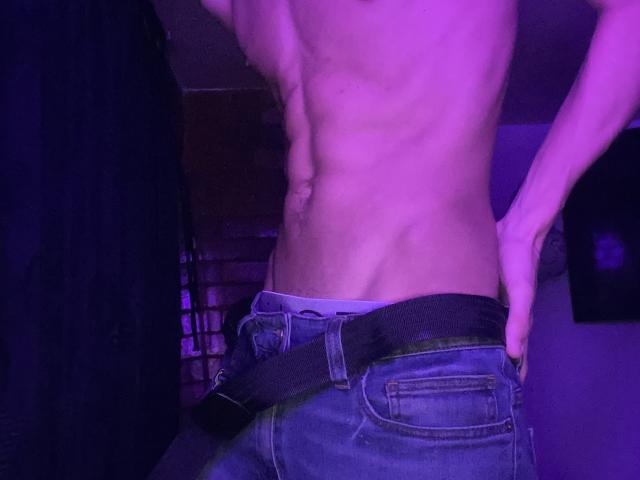 Angelitocute's bare, muscular back with defined abs and a slight waistline, wearing blue jeans with a black belt, in purple-lit room. Hands resting on hips.