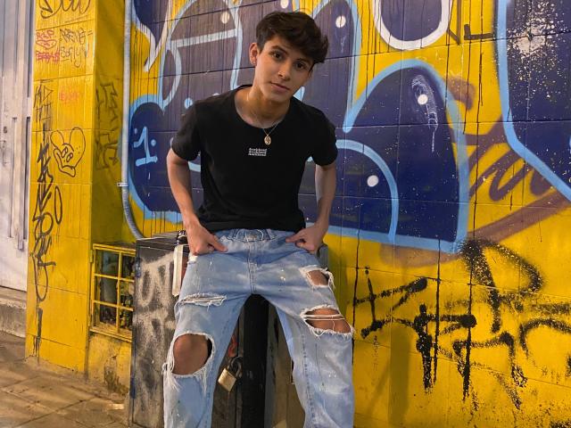 Angelitocute, young dude with dark hair, black tee, and ripped jeans, sitting on a trash can, hands on jeans, looking down, yellow and blue graffiti wall behind.