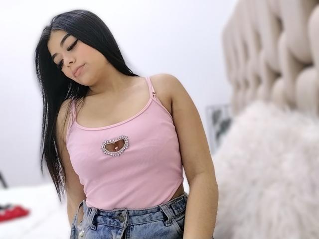 Ahnny with long black hair, wearing a pink tank top with a heart cutout, showing her tits, and blue jeans, standing in a bedroom. Her eyes are closed.