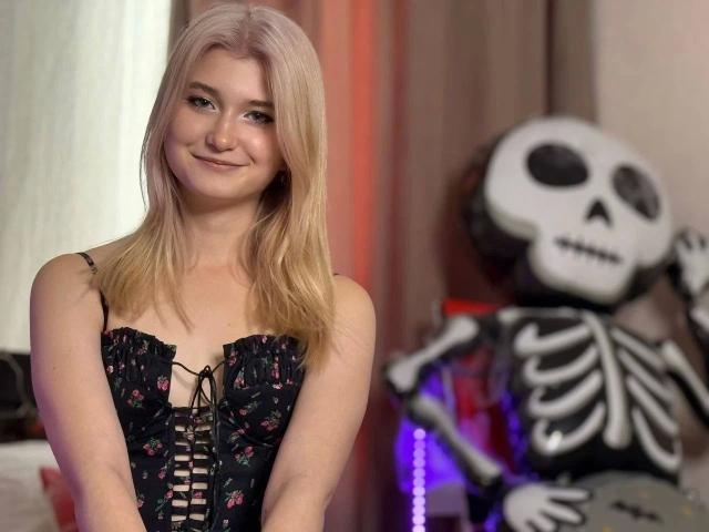 Monnyca-hot with blonde hair, smiling, wearing a black lace-up corset with pink floral patterns. Background has a large, white skull skeleton with black eyes, on the right. Monnyca-hot's corset is detailed with lace-up design, showcasing her fair skin. The skeleton's skeleton is illuminated with purple light. The overall composition contrasts Monnyca-hot's soft appearance with the stark, illuminated skeleton. The image uses a shallow depth of field, focusing on Monnyca-hot while the skeleton is slightly blurred.