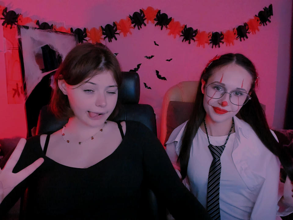 MonaNikol with small tits in black top, tongue out, next to MonaNikol with glasses, red face paint, white shirt, black tie, in Halloween-themed room. Red and pink lighting.