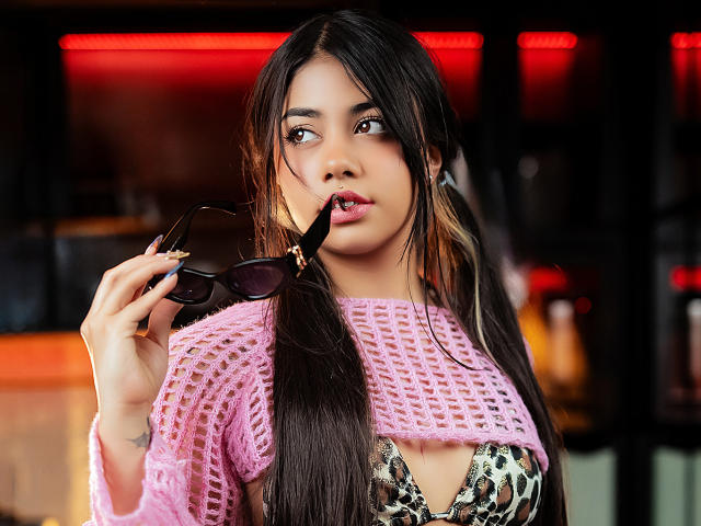 AnnyForger with long black hair, pink crochet top, and leopard print bra, holding black sunglasses near her pink lips, looking seductive, has a nose ring and tattoos.