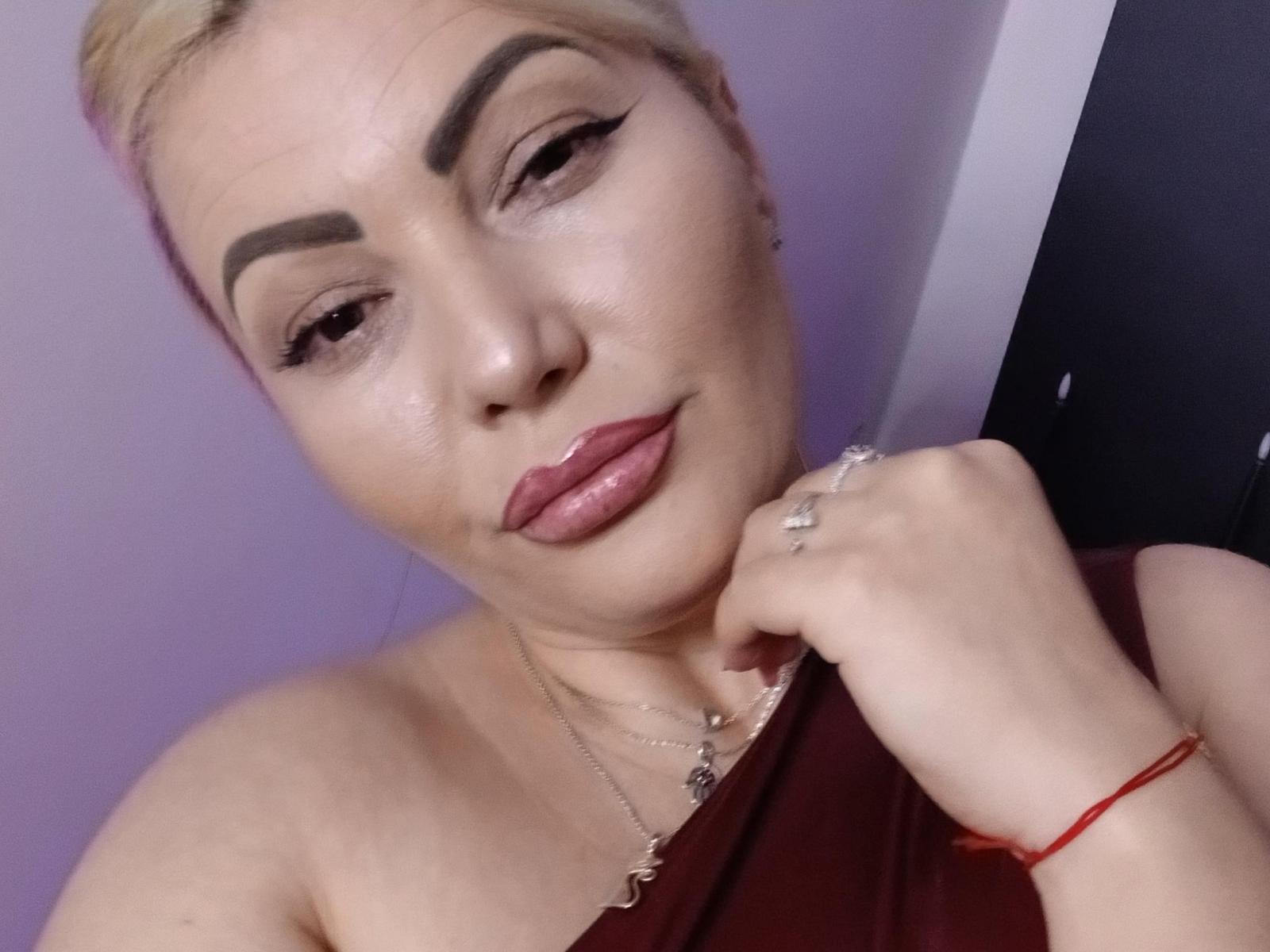 MistressDemi has light skin and blonde hair in a simple updo. She has thick, dark eyebrows, prominent eyeliner, and glossy pink lipstick. Her right hand, adorned with a silver ring and red wristband, touches her chin. She wears a one-shoulder maroon top and a delicate silver necklace. The image is a close-up selfie, showing her confident expression. The background is a soft purple wall.