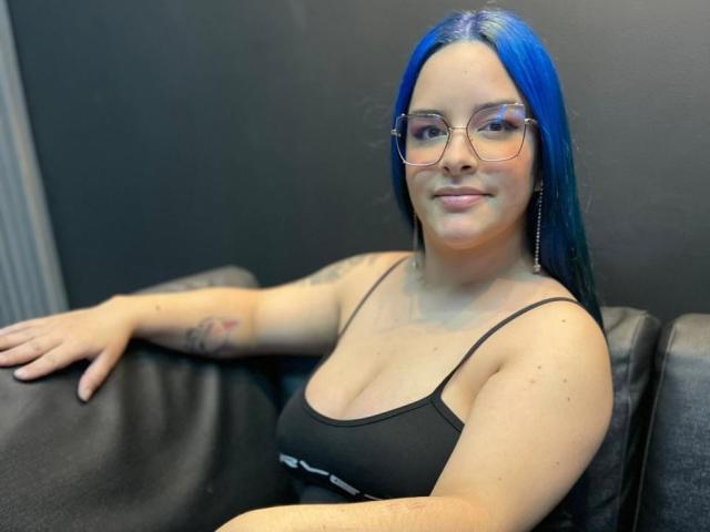 AuroraSantorini mit blauen Haaren, Brille, schwarzem Tanktop und Tätowierungen, sitzt auf einem schwarzen Ledersofa, lächelt und zeigt ihre kleinen Brüste und Armtätowierungen..
