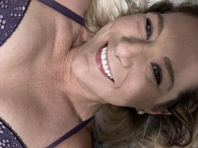 LilaJaneErotica with smiling face, white teeth, brown eyes, wavy blonde hair, purple lace bra, and small diamond earrings, lying down, showing a slightly wrinkled neck.