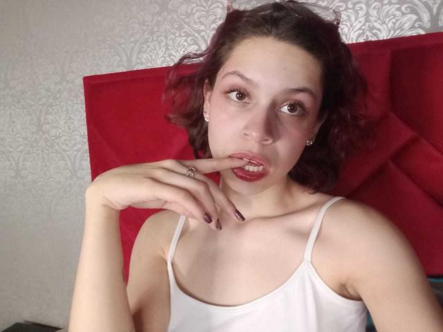 SophieBlake with red curly hair, pale skin, and pink lips, wearing a white tank top, touching her finger to her lip with purple nail polish, showing her tits.