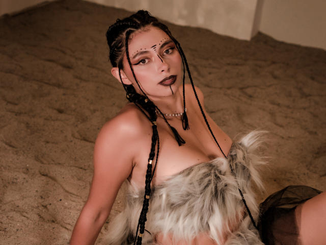 AnnaMooree, con cabello negro trenzado, lápiz labial oscuro y maquillaje en la cara, vestida con una prenda de piel que cubre sus senos, sentada en el suelo arenoso, mirando hacia arriba con una expresión seductora..