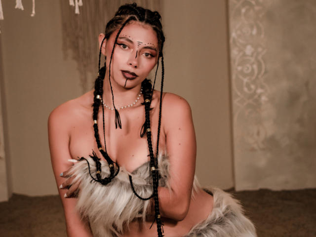 AnnaMooree, con cabello trenzado, lápiz labial oscuro y un collar de perlas, cubriendo sus senos con pieles, mirando seductoramente a la cámara. Su trasero es parcialmente visible..
